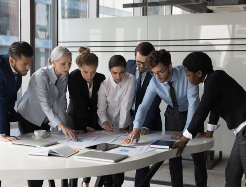 Diverse different aged business group standing at meeting table, negotiating on marketing reports, statistic diagrams, working on startup project together, using scrum approach with sticky notes